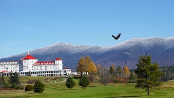 A large building with a red roof, surrounded by a green landscape and mountains in the background.