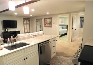A modern kitchen with granite countertops, a dishwasher, and a built-in oven. There is a dining area with a white table and chairs, and a living room with a sofa and a flat-screen TV. - Affordable Luxury! 4per. Efficiency Ski-out  Condo at Bretton Woods (Caroll)