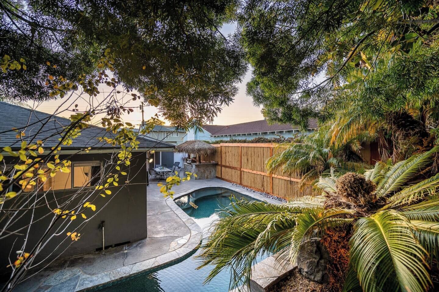 Un patio trasero con piscina, terraza de madera y una estructura con techo de paja.