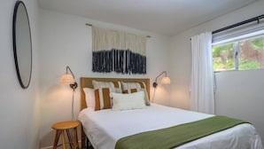 Una camera da letto con un letto, due abat-jour, un pannello decorativo in tessuto, uno sgabello rotondo in vimini e una finestra con tende.