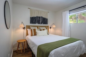 Ein Schlafzimmer mit einem Bett, zwei Nachttischlampen, einem gewebten Wandbehang, einem runden Korbstuhl und einem Fenster mit Vorhängen.