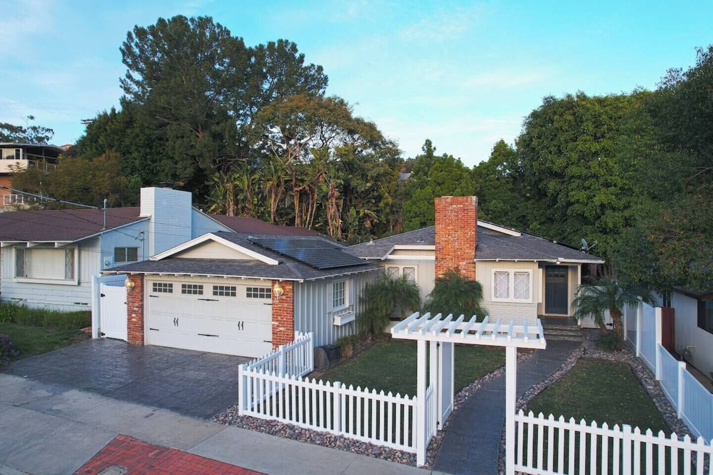 A residential area with two houses, each with a garage, surrounded by a white picket fence, and a small garden area with a pergola.