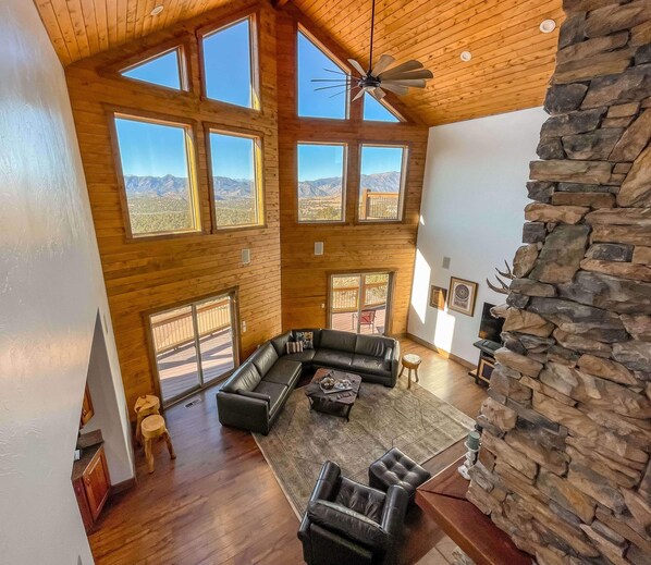 A spacious living area with a large L-shaped black leather sofa, a coffee table, and a stone fireplace. There are multiple windows offering views of the surrounding landscape.