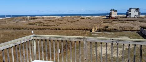 Une terrasse en bois donnant sur une plage bordée de dunes, avec un bâtiment visible au loin.