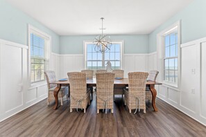 A dining room with a large table and wicker chairs, white walls, and a view of the ocean.