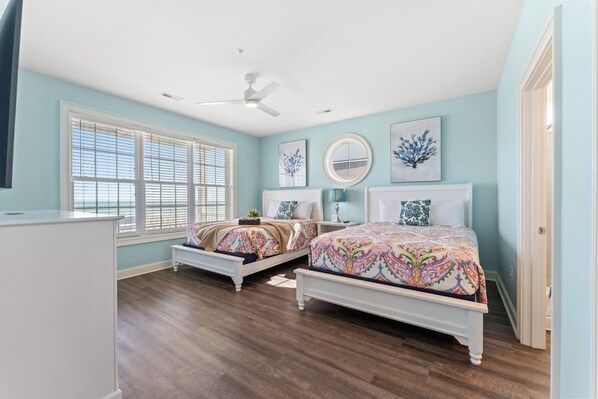 A bedroom with two beds, a ceiling fan, and large windows with blinds.