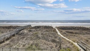 A beach with a wooden boardwalk leading to the shore, surrounded by dry grass and a clear sky.