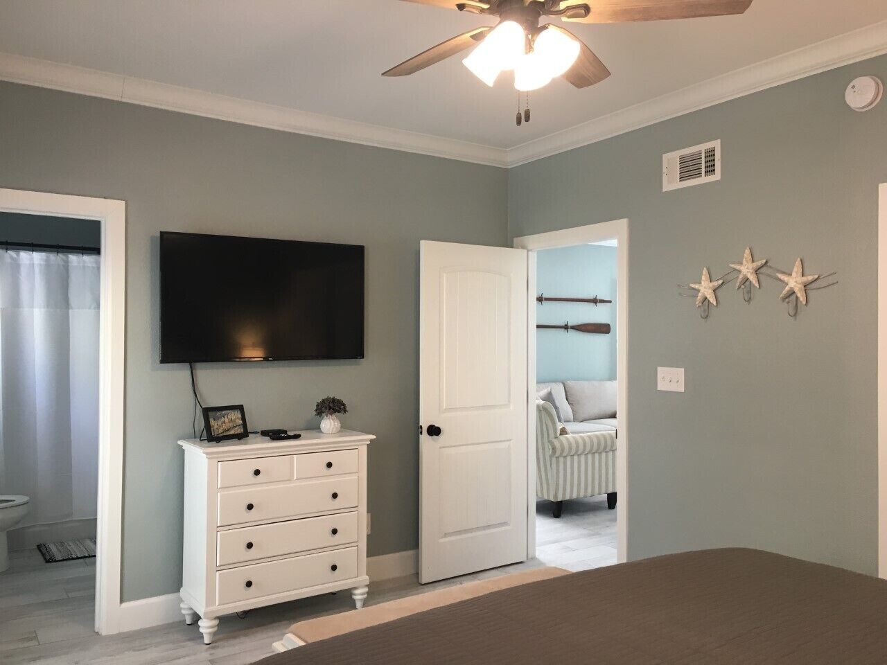 A bedroom with a ceiling fan, a TV mounted on the wall, a dresser, and a door leading to another room.