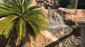 A waterfall cascading over rocks, surrounded by palm trees and a thatched-roof structure. - Slow Mo 17AD-Beautiful Beach Cottage @ the Purple Parrot Resort/Perdido Key FL (Pensacola)