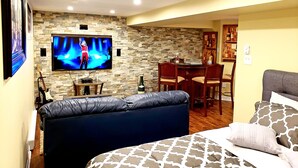 A cozy living room with a stone wall, a flat-screen TV, a leather sofa, a dining area with a table and chairs, and a bed with patterned pillows.