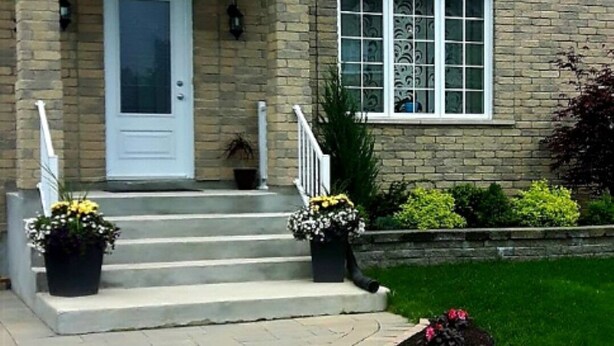 A two-story house with a front porch, a white door, and a small garden area with potted plants.