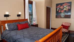 A neatly made bed with a grey quilt, a wooden headboard, and a red pillow. There is a small bedside table with a lamp and a vase of flowers. A chair with a red cushion is next to the bed. - Beautiful Home in Historic Neighborhood
1/2 mile to Downtown and the Riverwalk (san antonio)