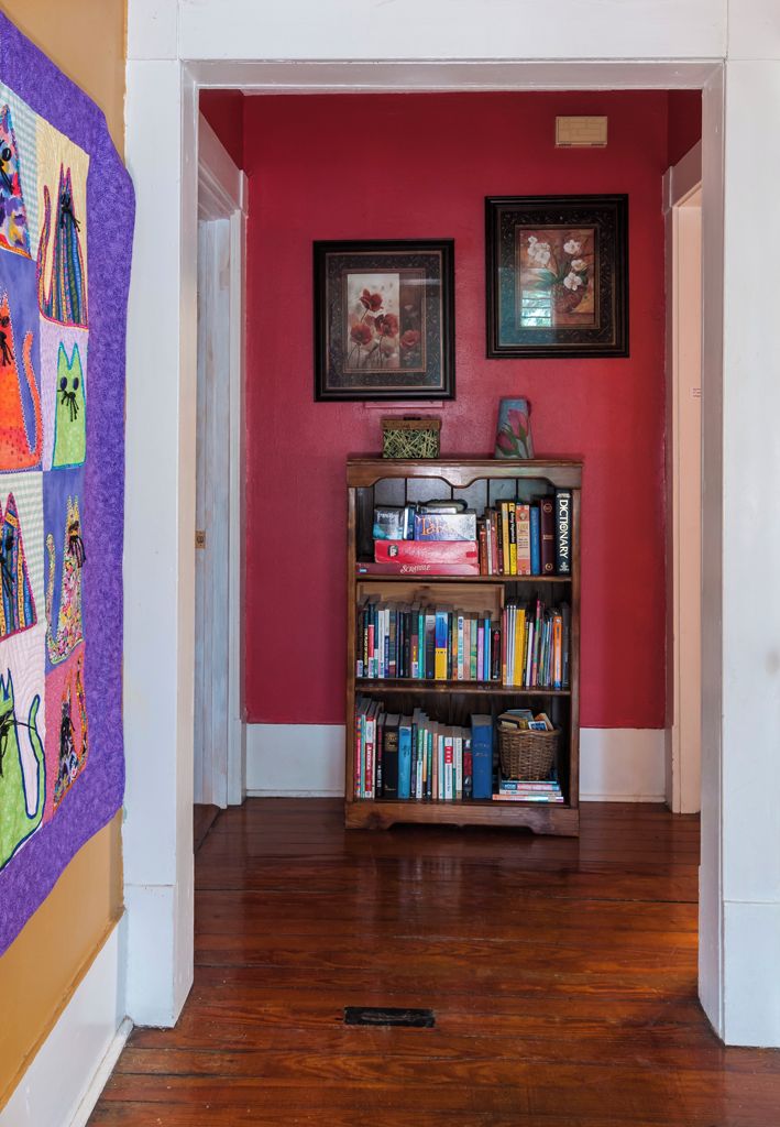 A hallway with a wooden bookshelf filled with books, a purple quilt on the wall, and two framed pictures above the bookshelf.
