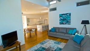 A modern living room with a grey sofa, a television, and a coffee table. There is a kitchen in the background with a dining area. - Husky Getaway - Villa with Heated Plunge Pool (Huskisson)