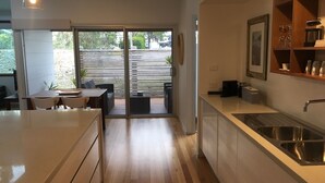 A modern kitchen with a large island, a dining area with a table and chairs, and a sliding glass door leading to an outdoor patio. - Husky Getaway - Villa with Heated Plunge Pool (Huskisson)