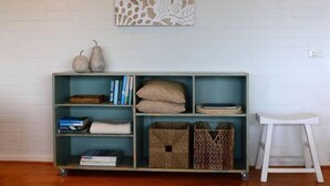 Una habitación con pared de ladrillo blanco, un estante azul con libros y objetos decorativos, además de un taburete blanco.