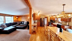 Un interior acogedor estilo cabaña, con detalles de madera, un sofá seccional, una mesa de comedor con sillas, una zona de cocina con electrodomésticos modernos y una lámpara de araña.