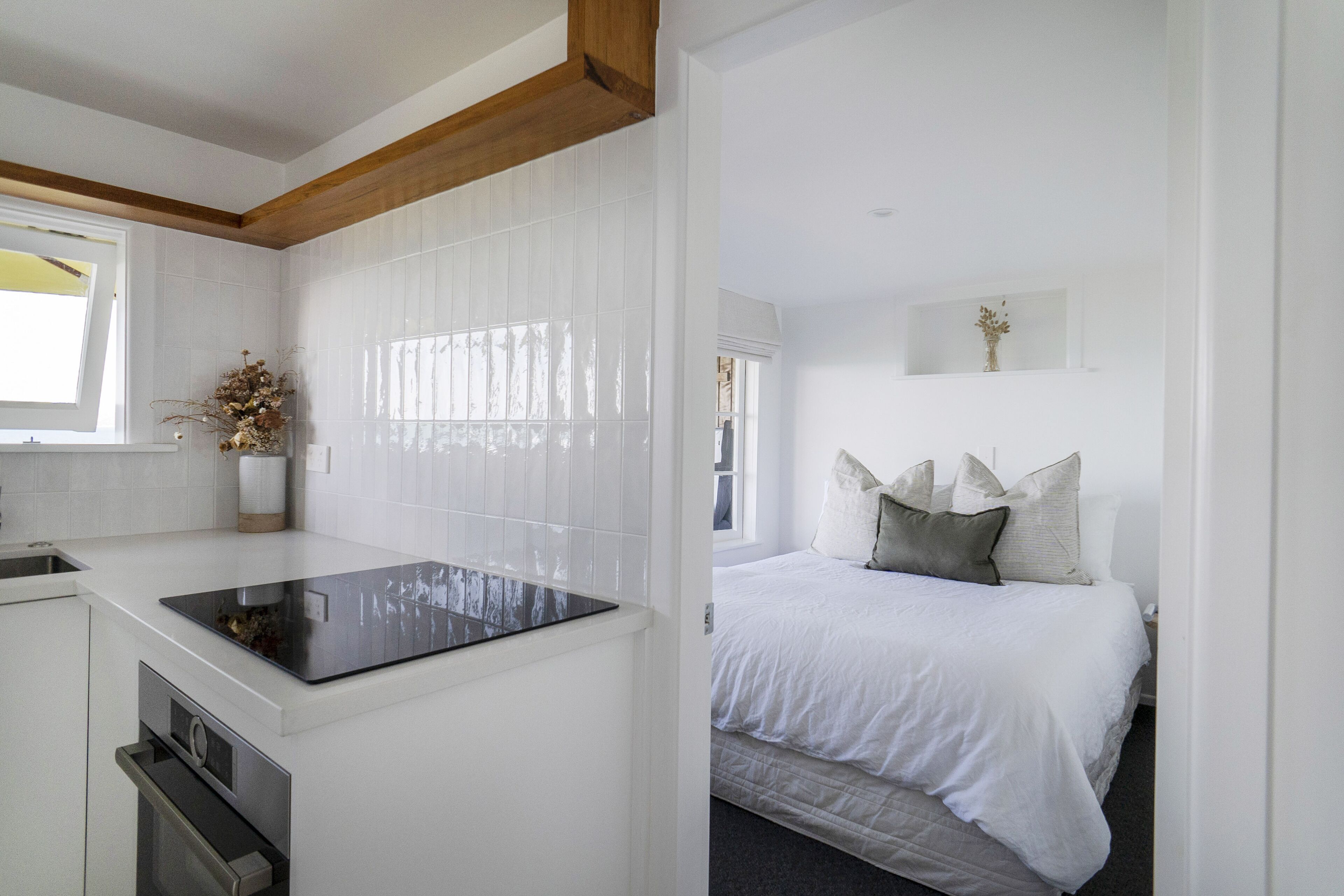 A modern kitchen with white cabinetry and a black countertop, a bedroom with a large bed and white bedding, and a window with blinds.