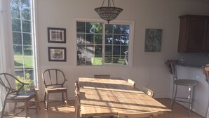 A dining area with a large wooden table and chairs, a hanging light fixture, and a view of a green landscape through large windows.
