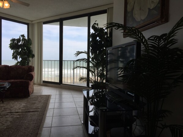 A living room with a large TV, a sofa, and a view of the beach through a sliding glass door.