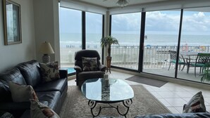 A living room with a glass coffee table, a sofa, and a chair. There is a view of the beach through large windows.