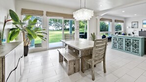 A spacious living area with a wooden dining table, a sofa, and a TV. There are large windows and a view of the outdoors. - 30 day Perfect “BEACHFRONT “ Getaway! Steps to the beach through the backyard (Laie)