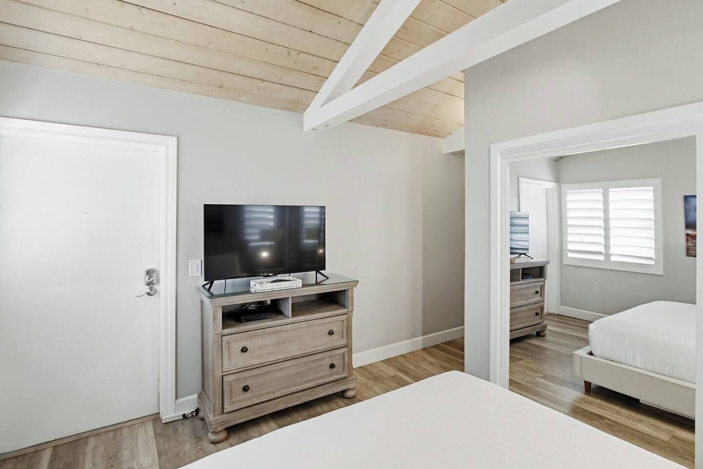 A bedroom with a TV on a wooden stand, a white bed, and a window with blinds.