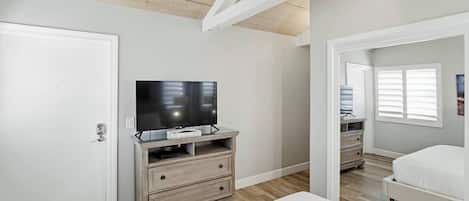 A bedroom with a TV on a wooden stand, a white bed, and a window with blinds.