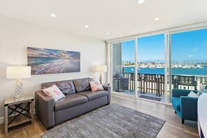 Un soggiorno moderno con un'ampia finestra che si affaccia su un porto turistico, un divano grigio con cuscini decorativi e un quadro raffigurante una spiaggia.