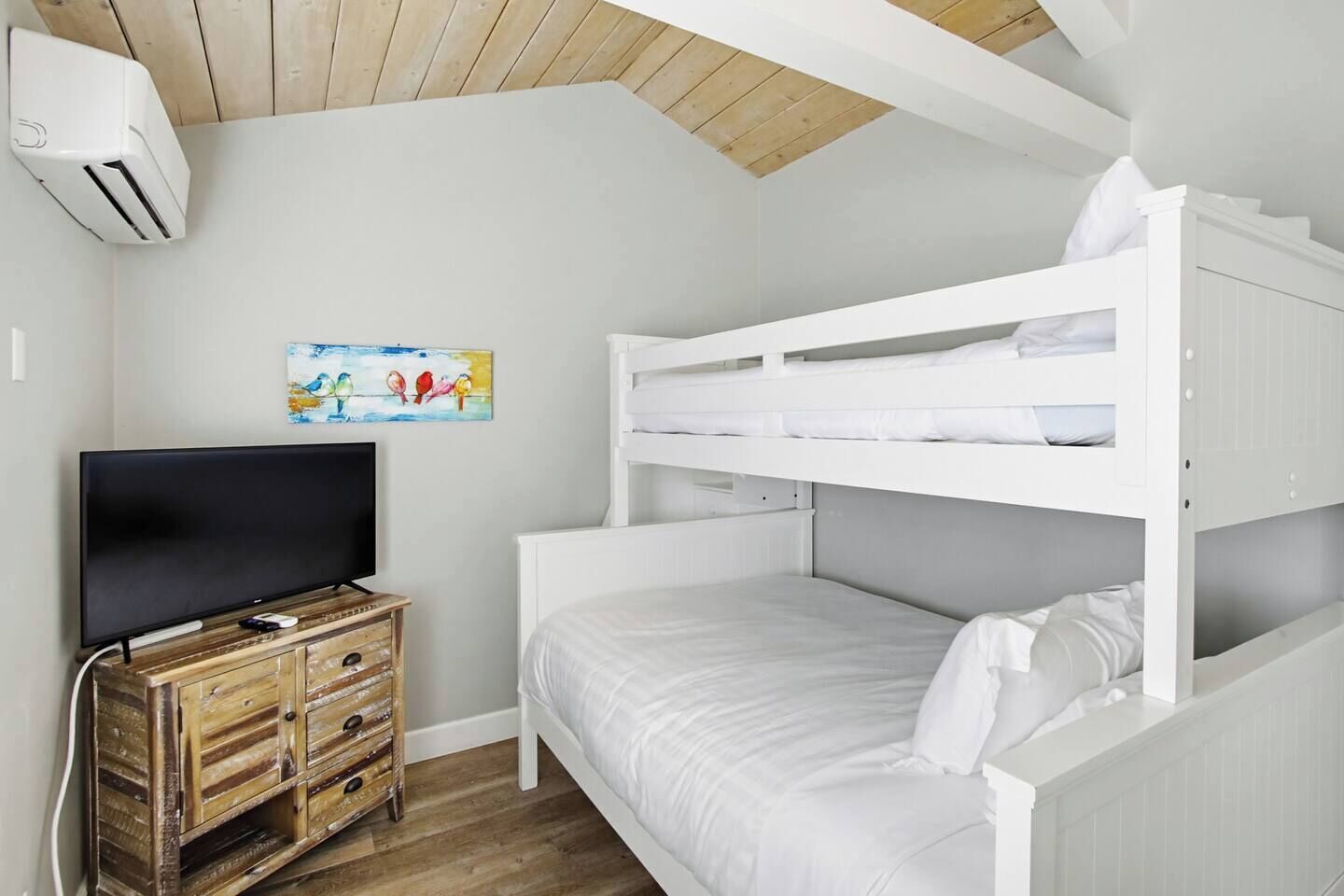 A bunk bed room with a television on a wooden dresser, white bedding, and a painting on the wall.