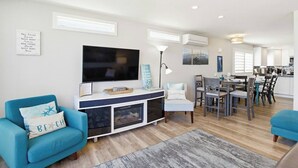 Ein modernes Wohnzimmer mit einem Flachbildfernseher, einem blauen Sofa, einem Essbereich mit Tisch und Stühlen sowie einer Küche im Hintergrund.