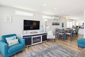 A modern living room with a flat-screen TV, a blue sofa, a dining area with a table and chairs, and a kitchen in the background.