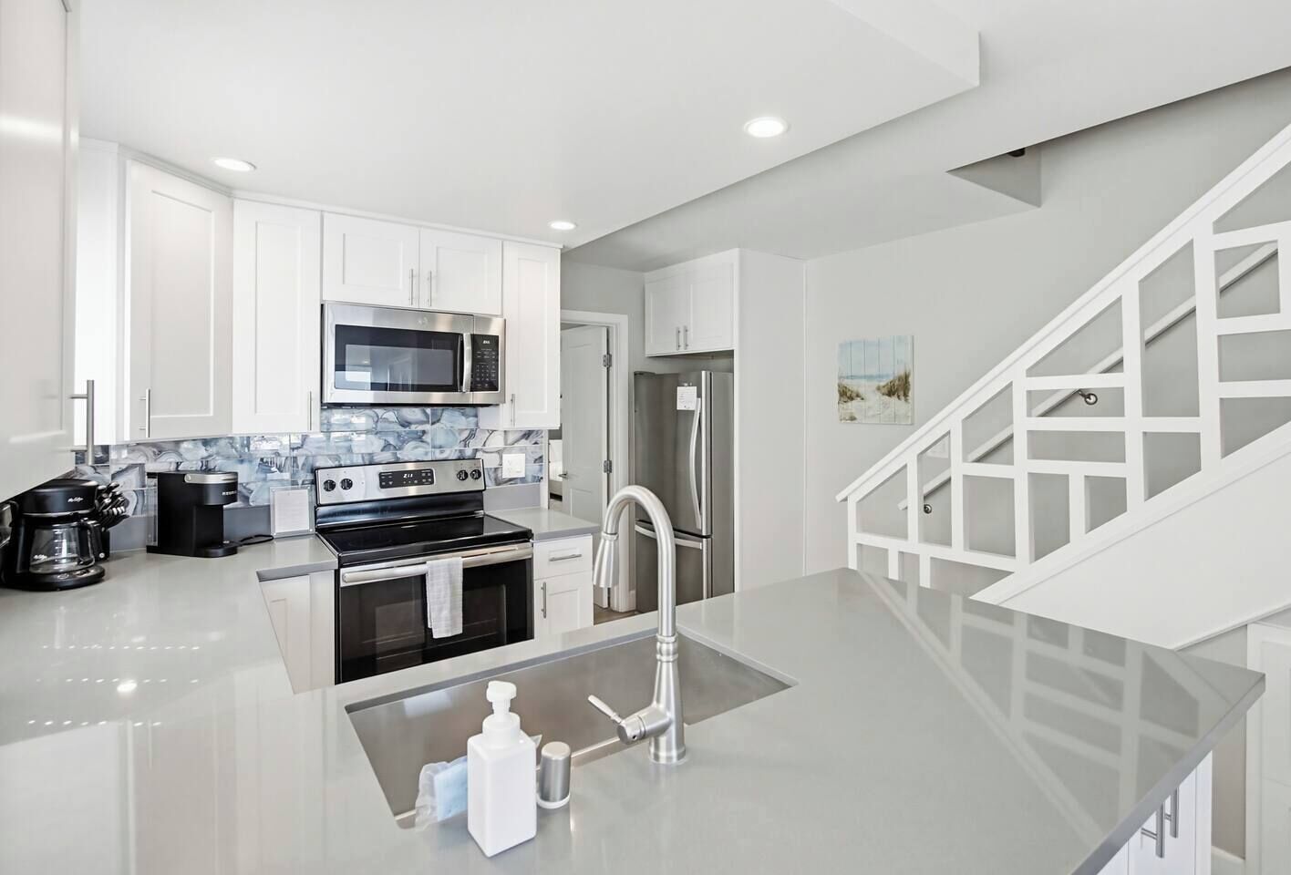 A modern kitchen with white cabinetry, a stainless steel refrigerator, a microwave, a coffee maker, and a sink with a soap dispenser.