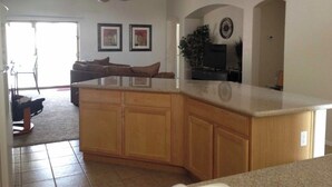 A spacious kitchen with wooden cabinets, a large island, and a ceiling fan. There are framed pictures on the wall and a plant on the counter. - Desert Escape Vacation Rental (Chandler)