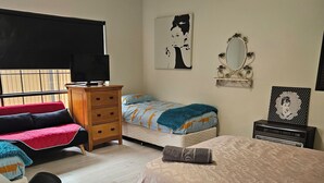 A bedroom with a bed, two bedside tables, a dresser, a desk, a chair, a TV, and a window with blinds.