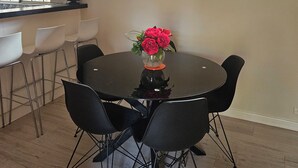A dining area with a round table, black chairs, and a vase of red flowers. There is a window with a brick wall outside, and a bookshelf with books and decorative items.