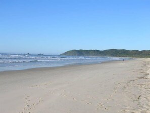 Une vaste plage de sable fin, avec des vagues douces, un ciel dégagé et une colline boisée en arrière-plan.