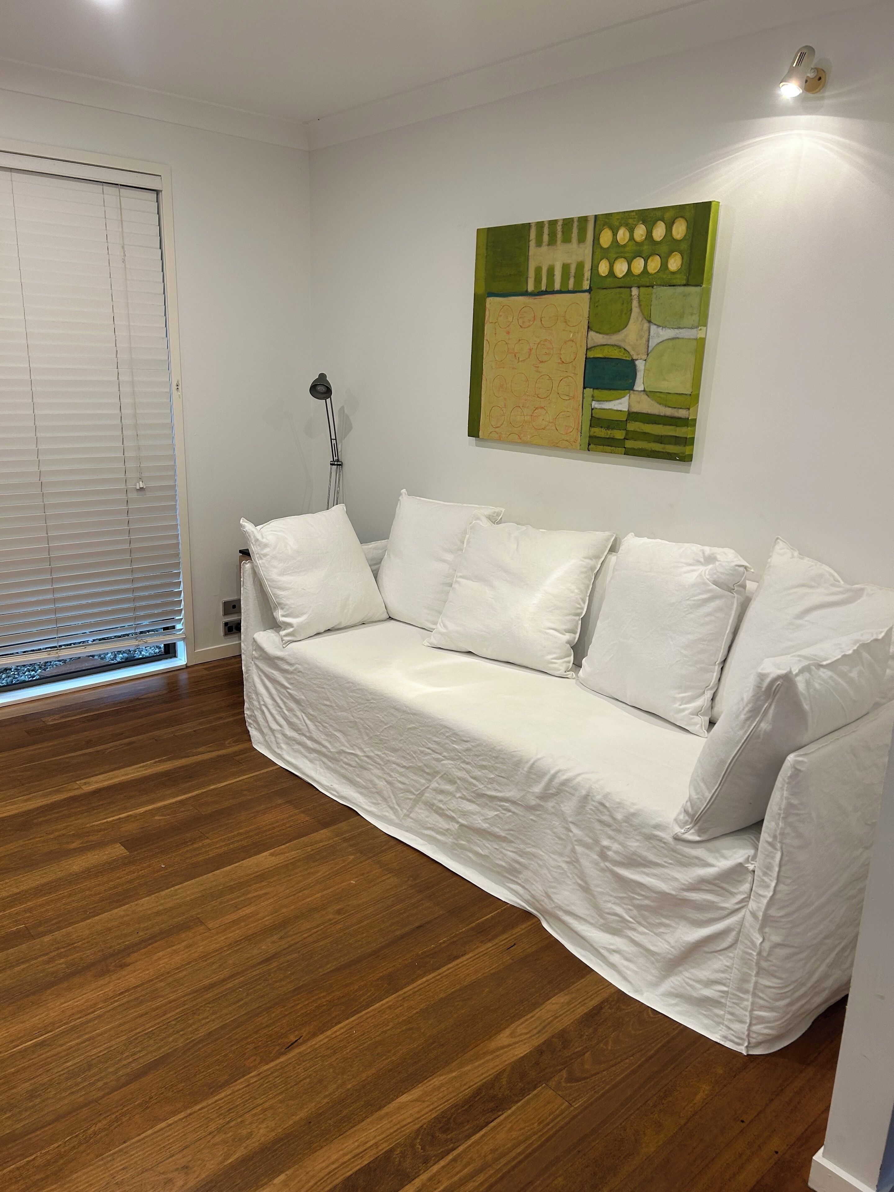 Un canapé blanc avec des coussins, un lampadaire et un tableau abstrait vert accroché au mur.