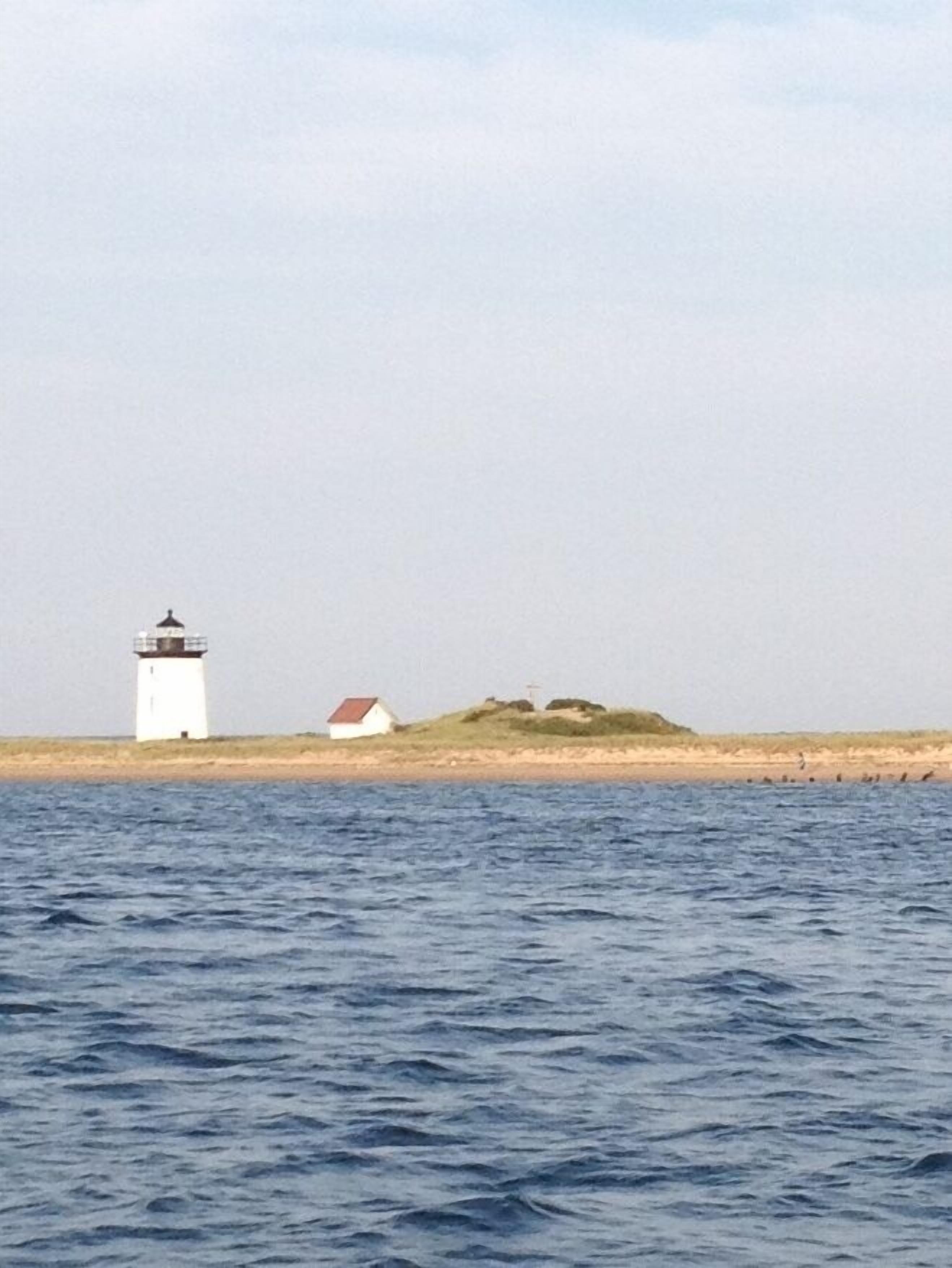 Ein Leuchtturm und ein kleines Gebäude an einem sandigen Ufer mit ruhigem Wasser im Vordergrund.