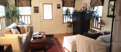 A living room with a sofa, ottoman, and a red patterned rug. There is a door, windows with curtains, and a plant.