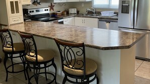 Cocina moderna con armarios blancos, encimera de granito, refrigerador de acero inoxidable y una isla central con taburetes de bar.