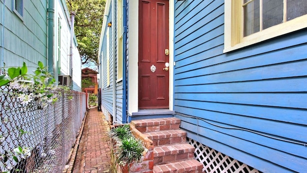 A narrow alleyway between two blue houses with a red door, brick steps, and a chain-link fence with plants. - Camelback Guest House (New Orleans)