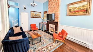 A living room with a blue wall, a brick fireplace, a TV, and a large painting. - Camelback Guest House (New Orleans)