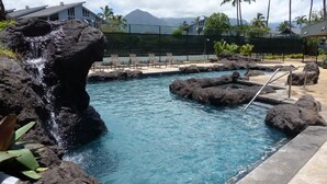 A pool with a rocky edge and a waterfall feature, surrounded by lounge chairs and palm trees. - Prime Oceanfront End Unit--View--Family-friendly Cliffs Resort at Bargain Rates (Princeville)