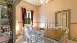 A dining room with a wooden table and white chairs, a chandelier, and a view through a window with curtains. - Historic 1885 Blue Mountains Retreat | Sleeps 12 | 6 Bdrms | Free EV Charging (Wentworth Falls)