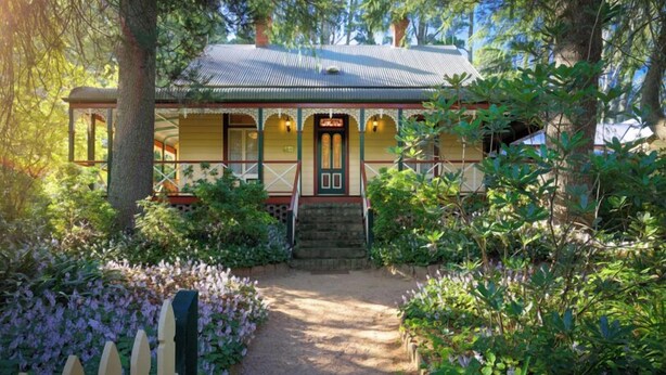 A traditional building with a covered porch, surrounded by lush greenery and a gravel path leading to the entrance. - Historic 1885 Blue Mountains Retreat | Sleeps 12 | 6 Bdrms | Free EV Charging (Wentworth Falls)