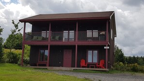 Un immeuble de deux étages avec un balcon à l’étage supérieur, une porte d’entrée et deux chaises rouges à l’extérieur.