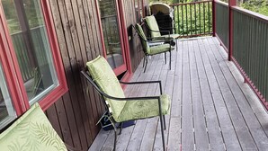 Une terrasse en bois avec deux chaises vertes dotées de coussins et une table, une rambarde rouge et une vue sur les arbres.