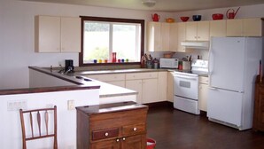 Une cuisine avec une grande fenêtre, des armoires blanches, une chaise en bois et un réfrigérateur.