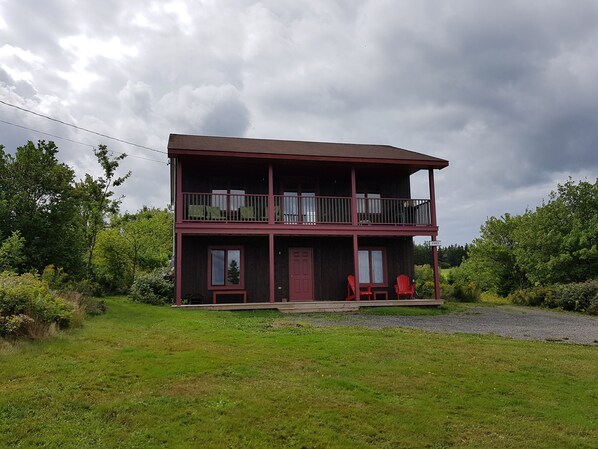 A two-story wooden house with a balcony, a front porch with red chairs, and a grassy yard. - Year-round Vacation Rental - Fly Fishing, Golf, Beaches, Cabot Trail & More.
 (Margaree)